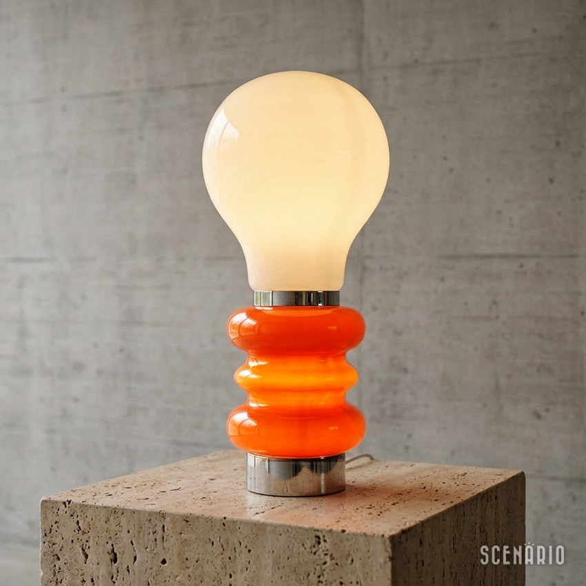 Lampada-da-tavolo-Space-Age-accesa-piedistallo-travertino.jpg Fotografia di una lampada da tavolo accesa in stile Space Age anni '70, caratterizzata da un grande bulbo superiore in vetro bianco opalino e una base sagomata a onde in vetro arancione acceso. La lampada poggia su un piedistallo quadrato in travertino beige poroso, sullo sfondo di una parete di cemento a vista. L'immagine ha un'atmosfera retrò e moderna.