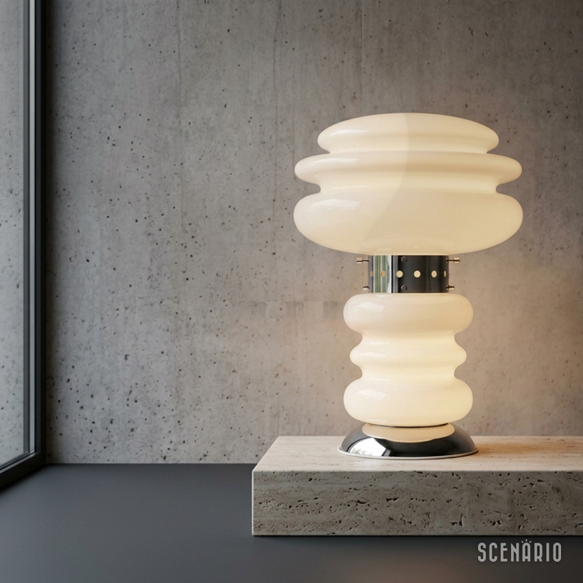 Lampada-Mazzega-Space-Age-vetro-opalino-travertino.jpg Fotografia d'interni con lampada Space Age attribuita a Carlo Nason per Mazzega, accesa. Il diffusore e la base sagomata in vetro opalino bianco sono uniti da una fascia centrale in cromo perforato. La lampada poggia su un piedistallo basso in travertino beige poroso, poggiato su un pavimento in ardesia scura. Lo sfondo è un muro di cemento a vista. Sulla sinistra, una grande finestra con cornice nera. La luce emessa è calda e diffusa.